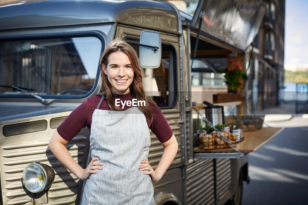 Portrait of smiling female owner standing hands on hips outside food truck in city