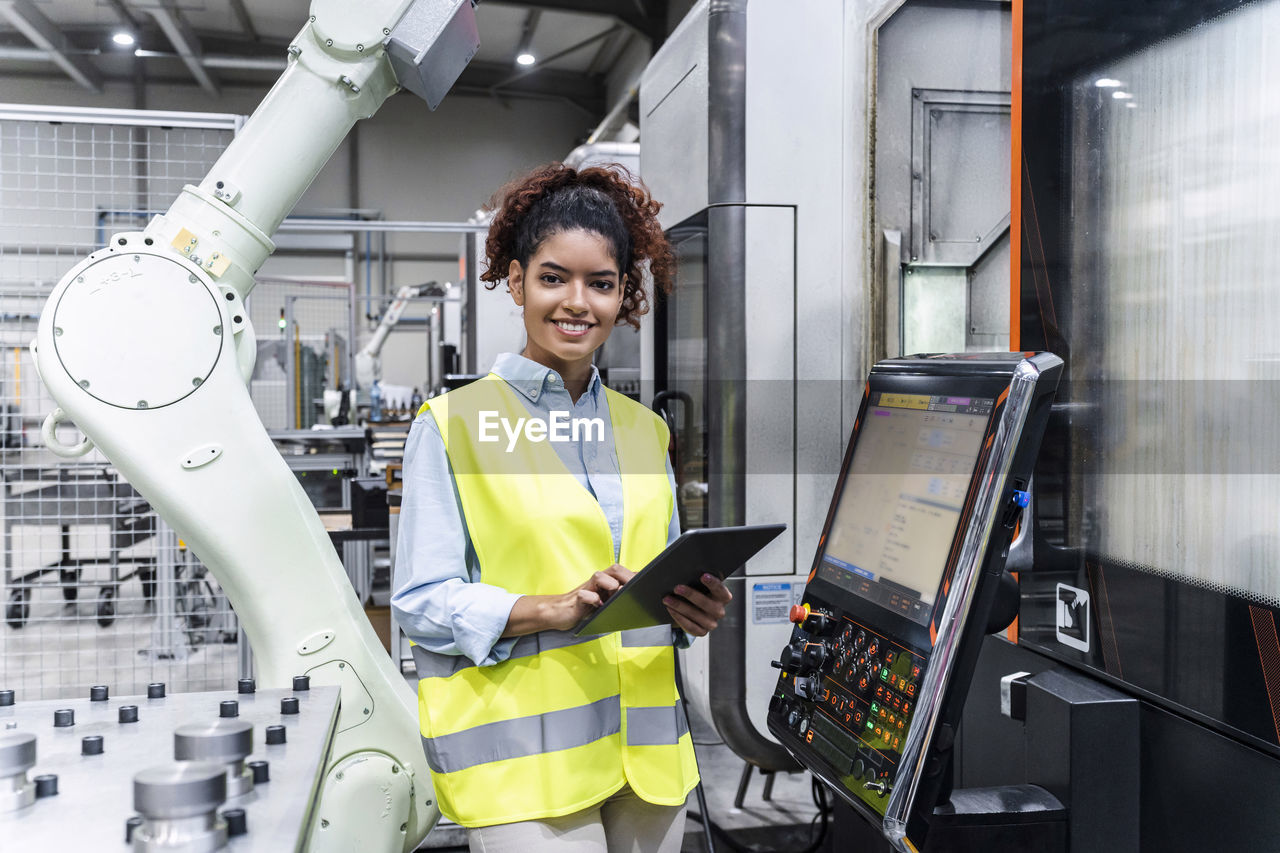 Happy engineer standing with tablet computer by machine in factory