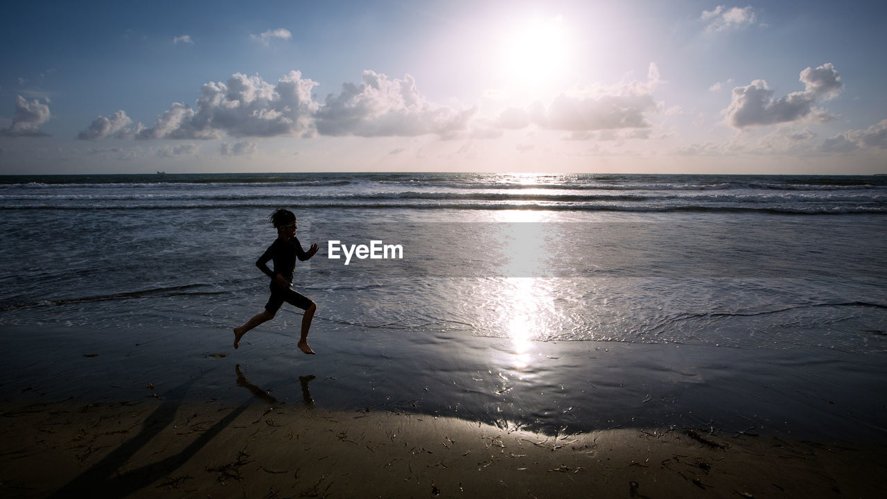 Side view silhouette of boy running on beach against sky