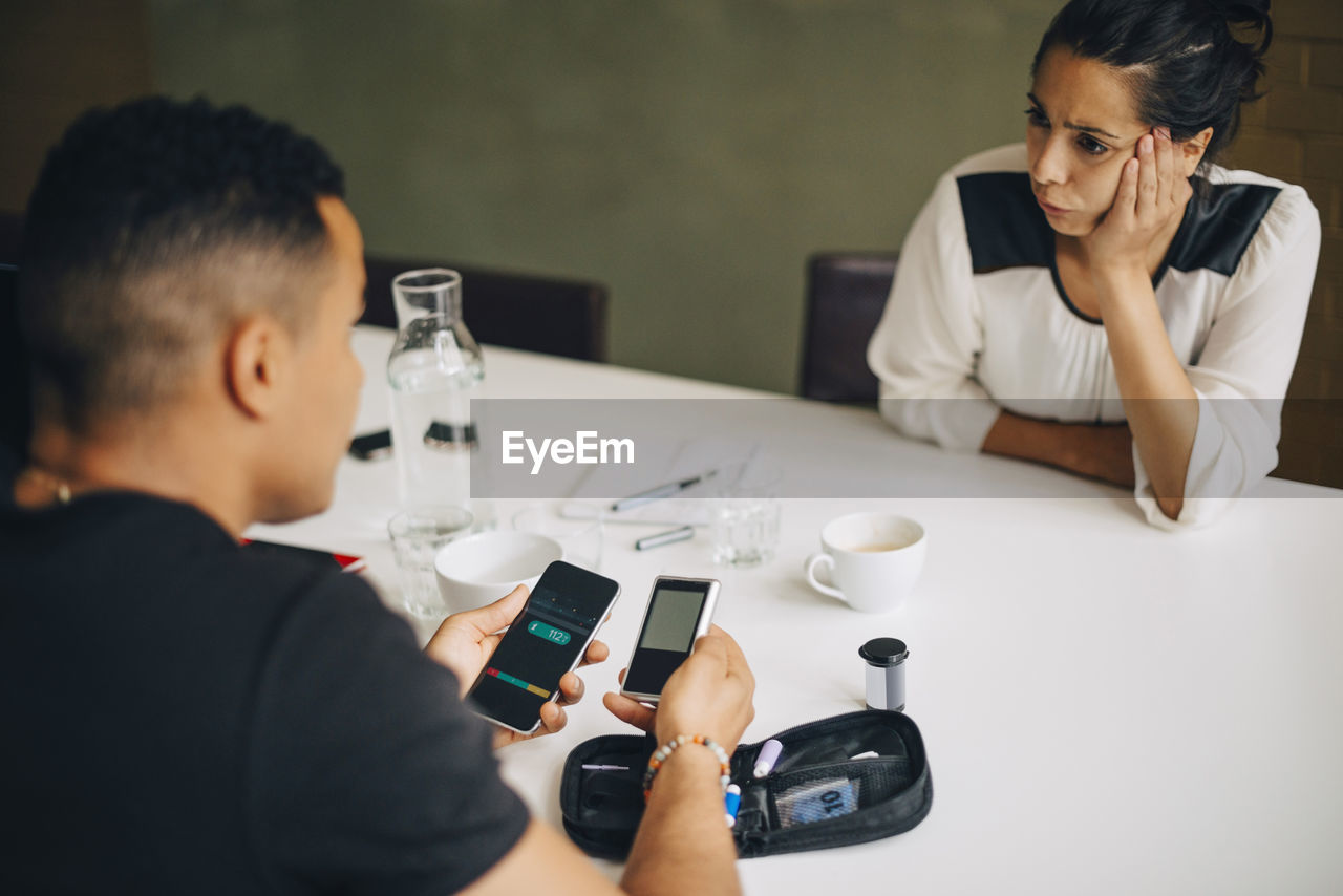 Colleague looking at businessman doing blood sugar test while sitting at table during coffee break