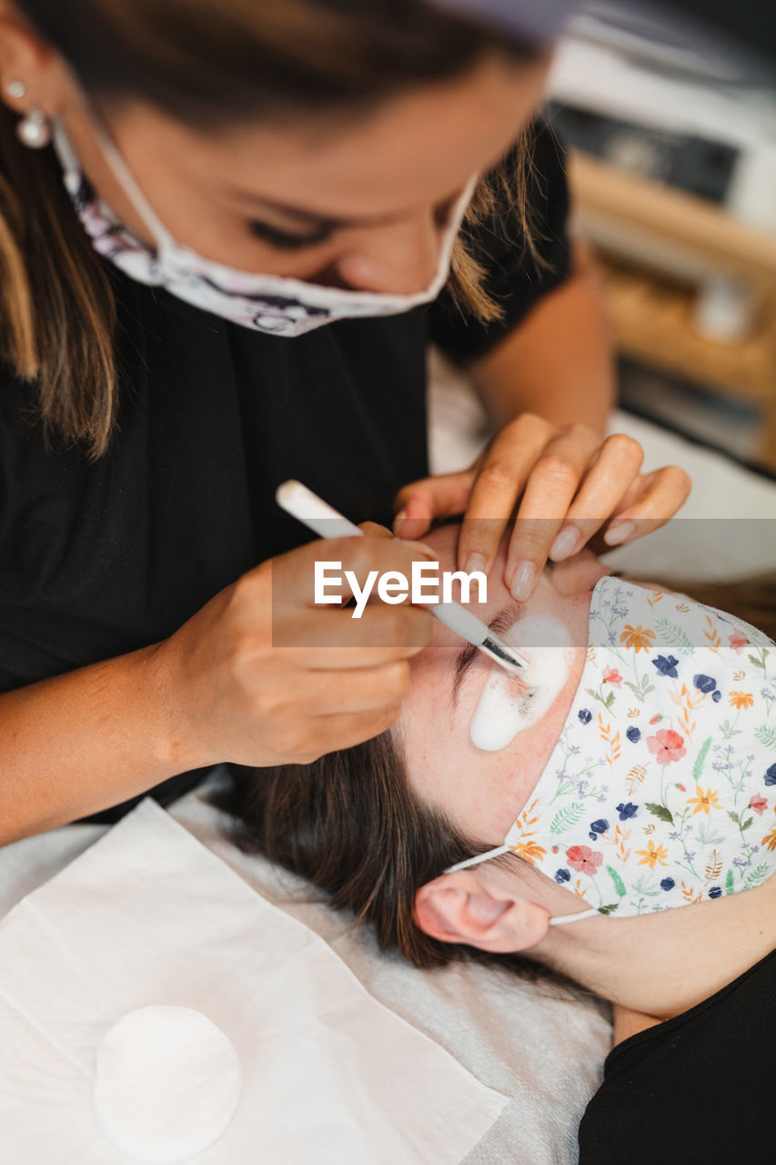 High angle of crop anonymous female master in medical mask applying foaming cleanser on eyelashes of client after extension in modern salon