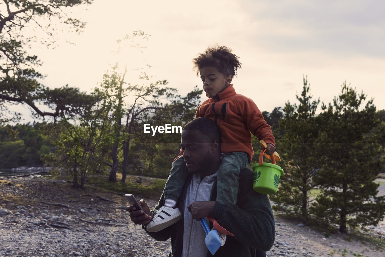 Father using mobile phone while carrying son on shoulders against sky