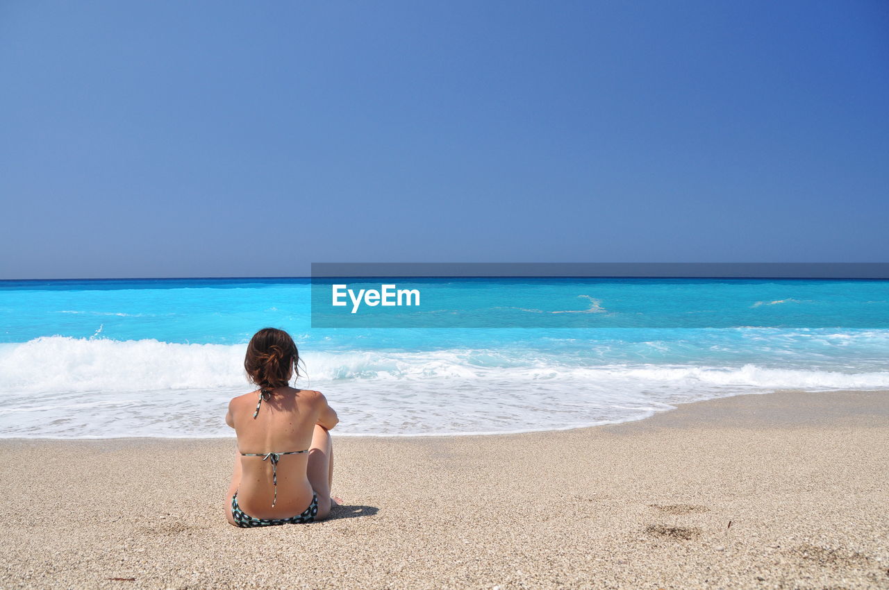 Rear view of woman in bikini sitting at sandy beach against clear sky