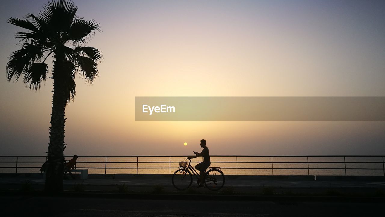 SILHOUETTE PEOPLE RIDING BICYCLE ON SHORE AGAINST SKY DURING SUNSET