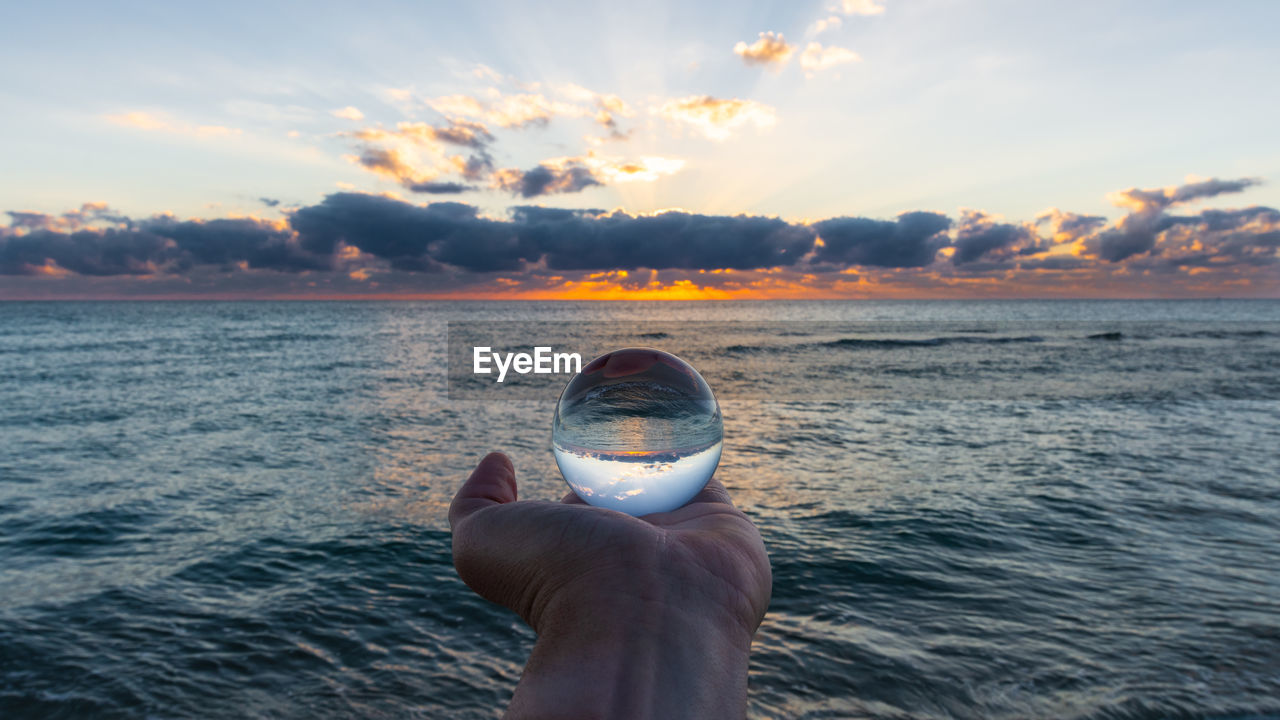 Cropped hand person holding crystal ball by sea against sky during sunset