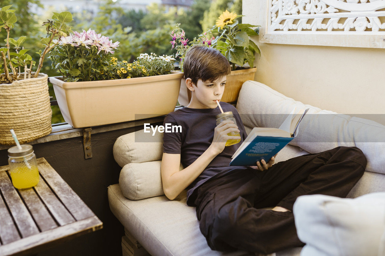 Boy drinking juice and reading book while reclining on sofa in balcony