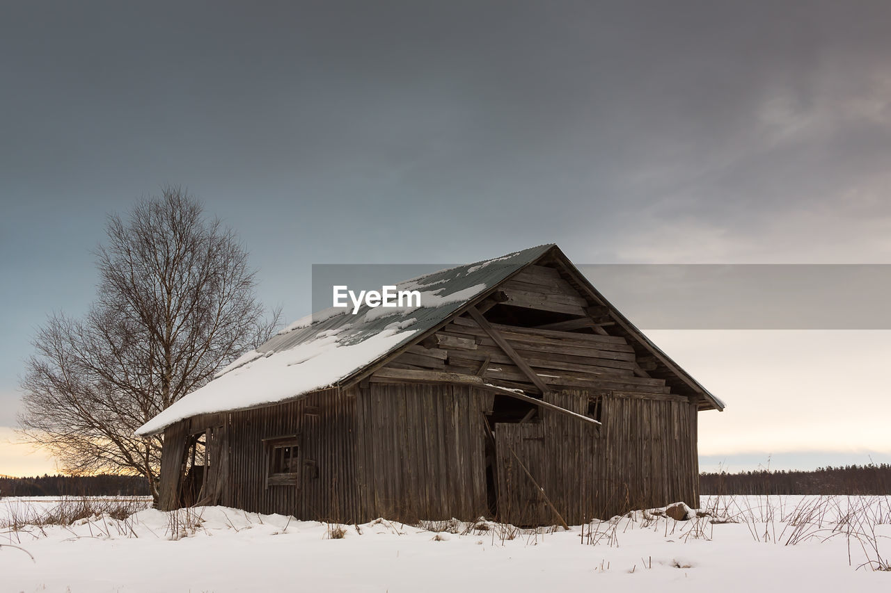 Abandoned house on snow covered field against cloudy sky