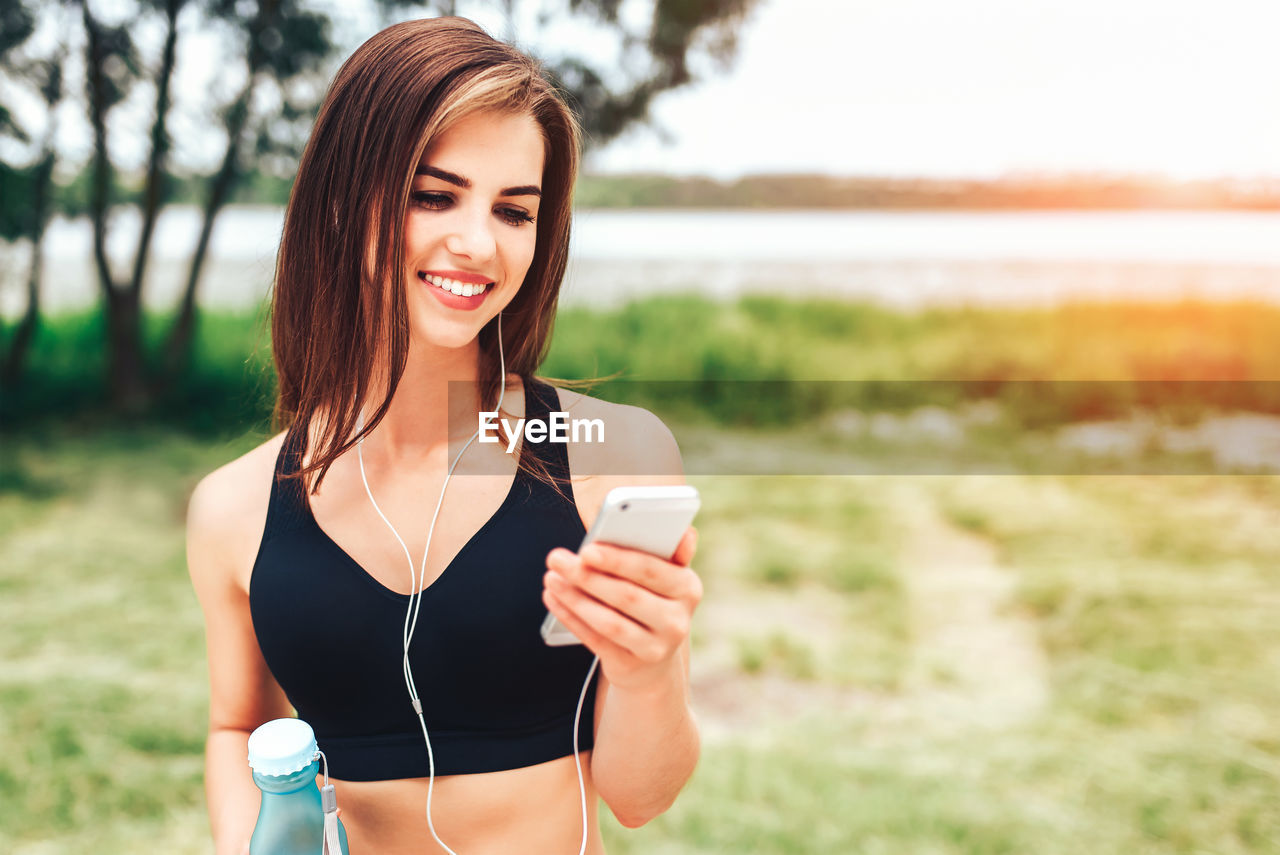 Smiling young woman listening to music from mobile phone while standing on field