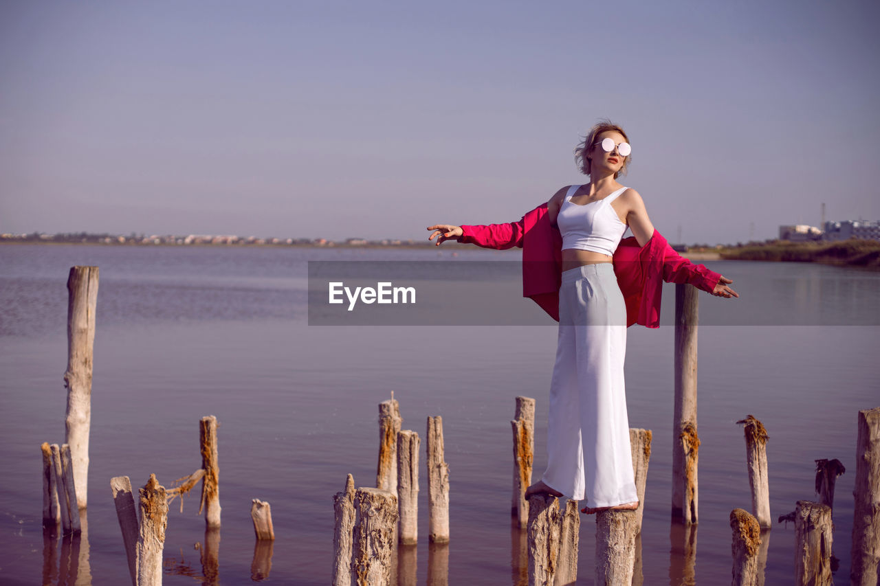 Portrait of young woman in pink and white clothes wearing sunglasses stands on stump on pink lake 