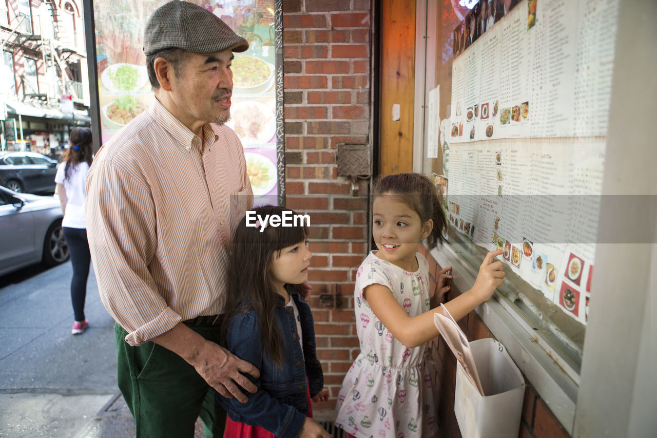 Girl showing menu to grandfather and sister while standing against restaurant
