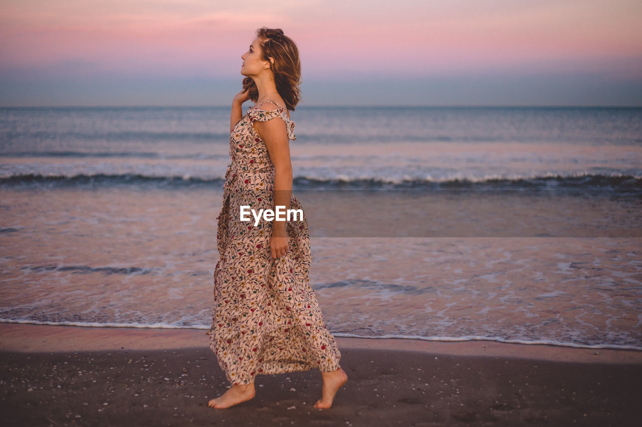 Young woman walking at beach against sky during sunset