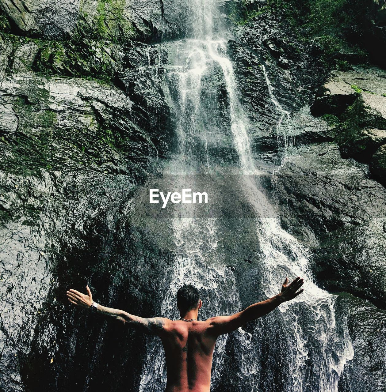 Rear view of shirtless man standing towards waterfall