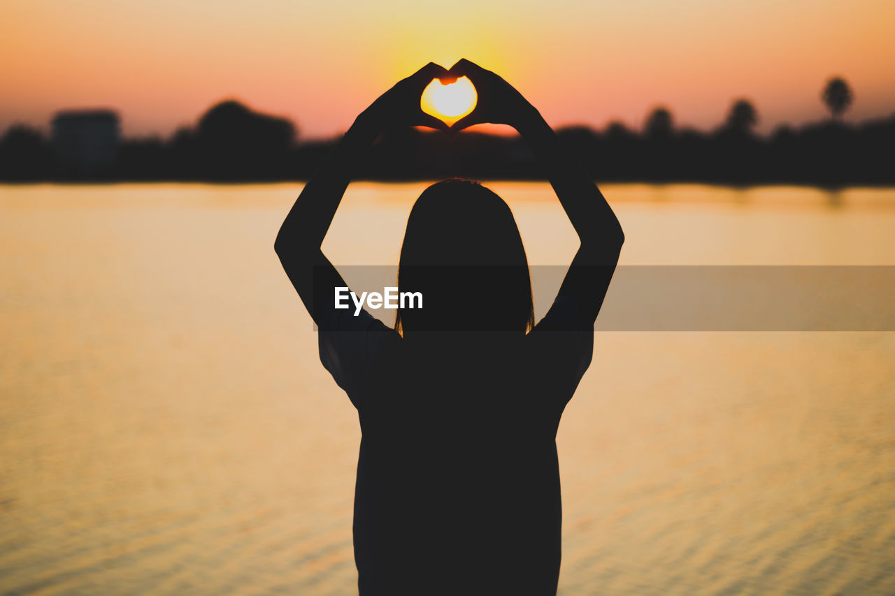 Silhouette woman making heart shape with hands by lake against sky during sunset