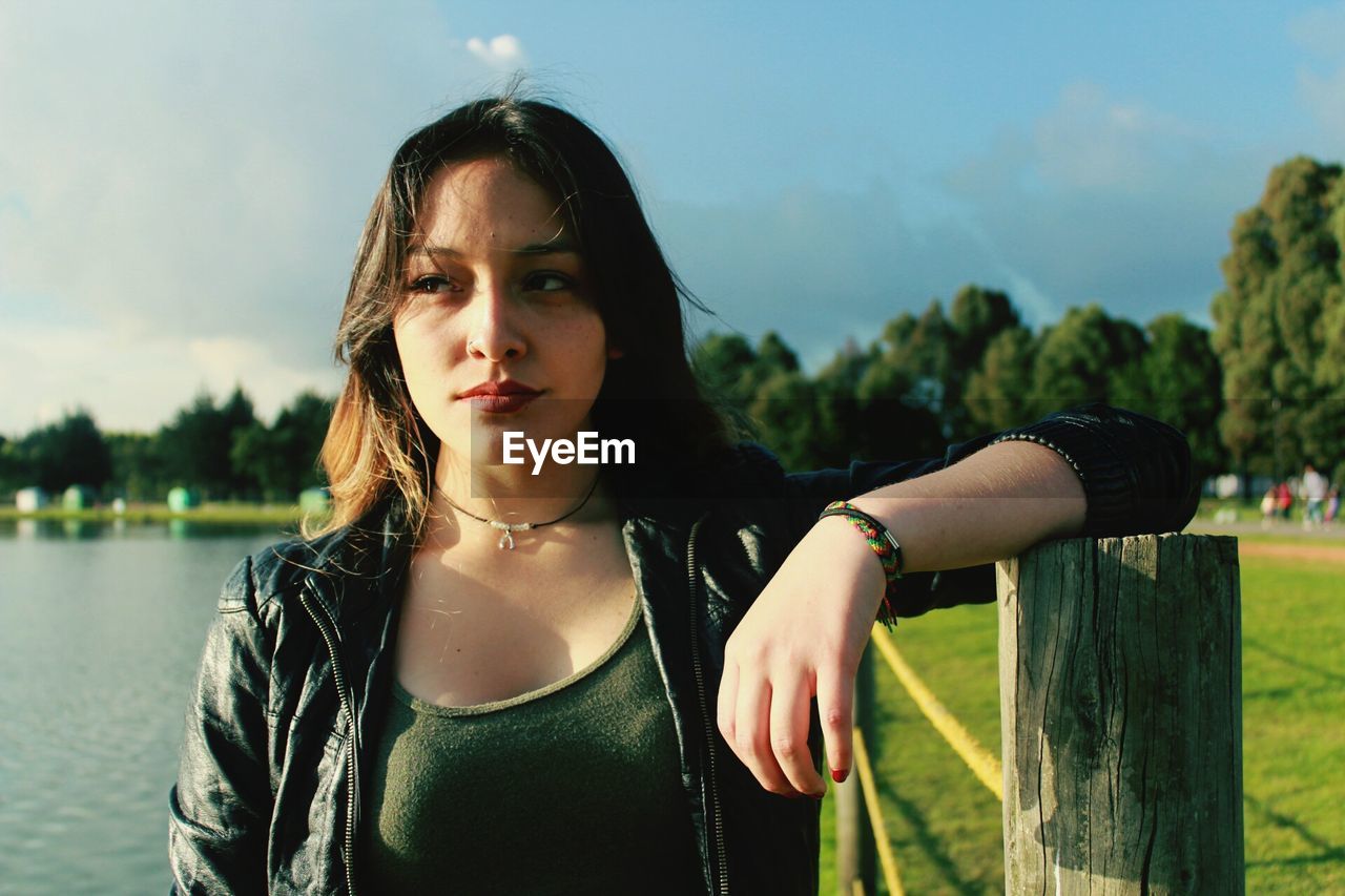 Thoughtful young woman looking away while standing by wooden post at park