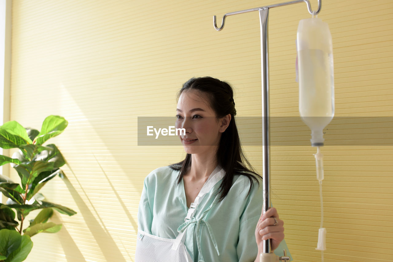 Smiling woman with iv drip standing against wall