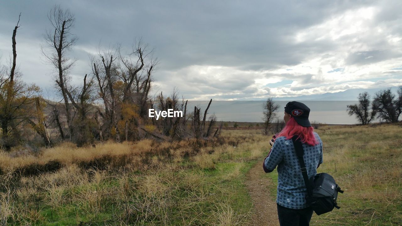 REAR VIEW OF WOMAN STANDING ON FIELD AGAINST CLOUDY SKY