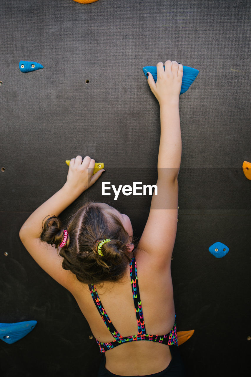 Back view of anonymous child in sportswear climbing wall with artificial rocks while training in gym and enjoying weekend