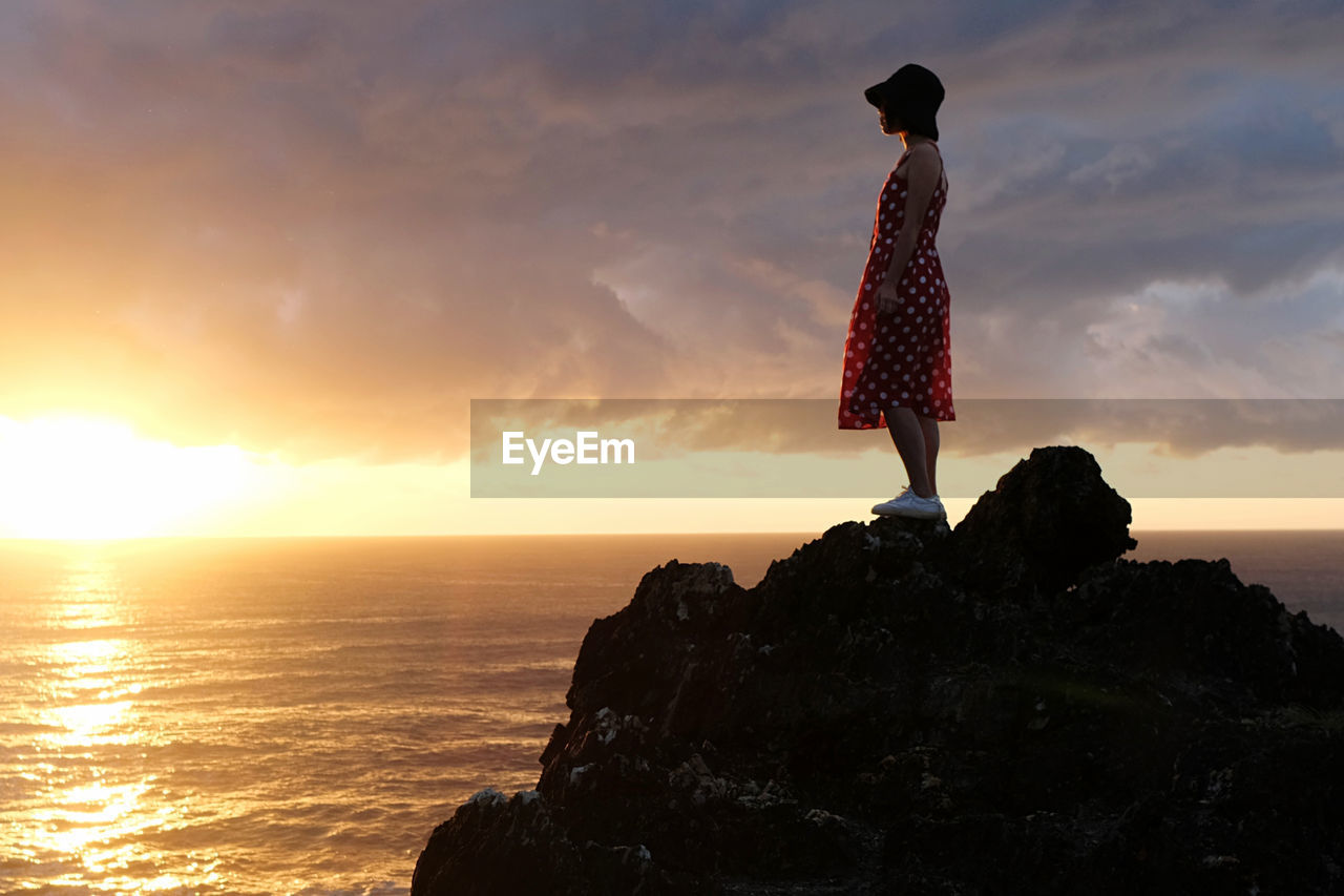 rear view of woman standing on rock against sky during sunset