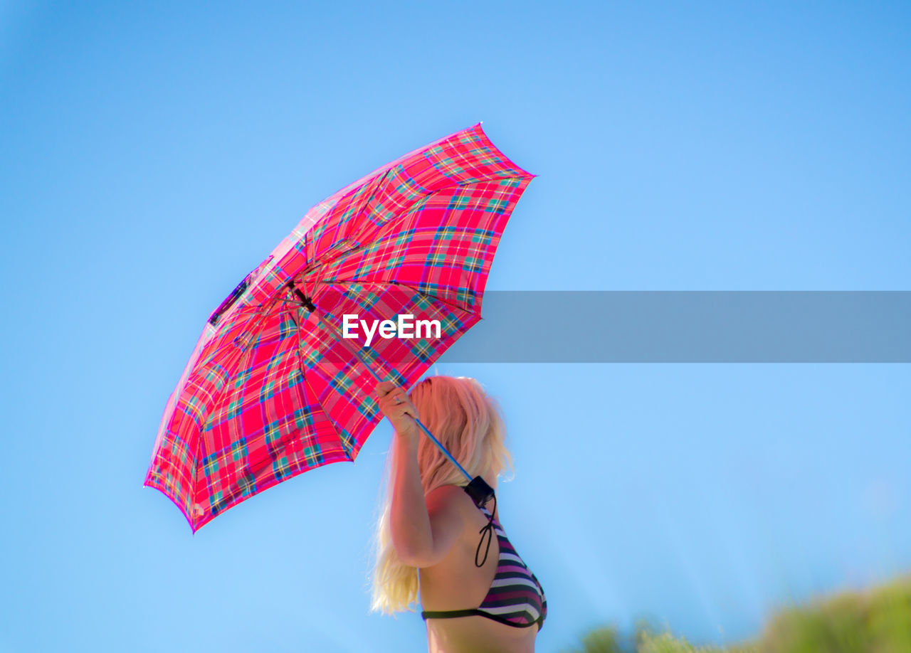 Side view of woman holding umbrella while standing against clear blue sky