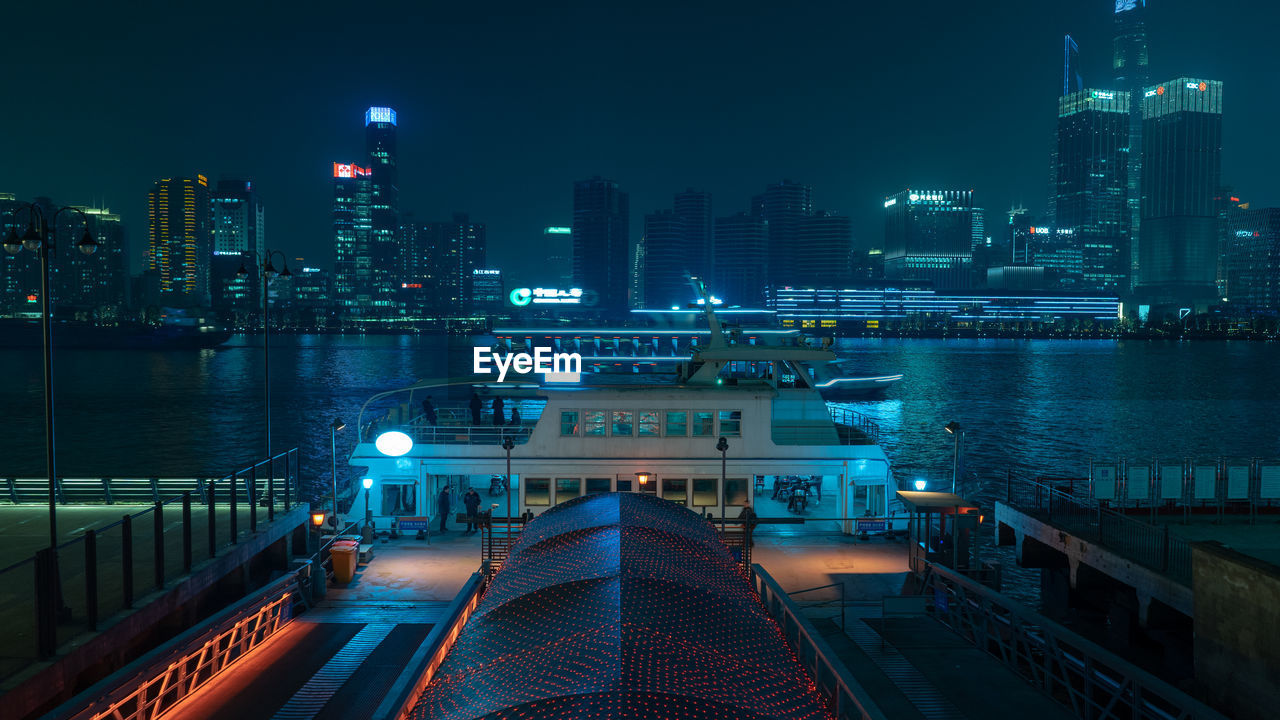 Illuminated modern buildings by river against sky in city at night