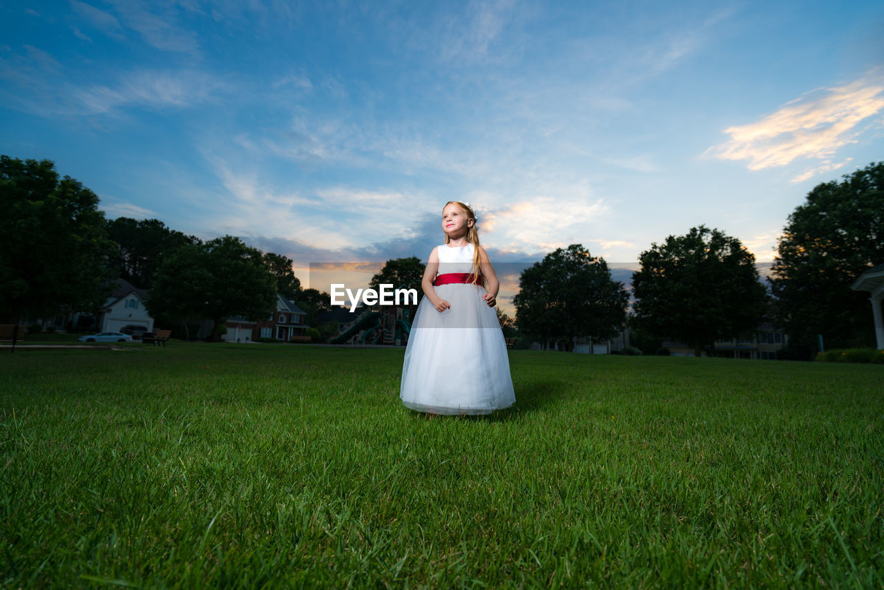 Cute flower girl in white dress standing on grassy field against sky during sunset