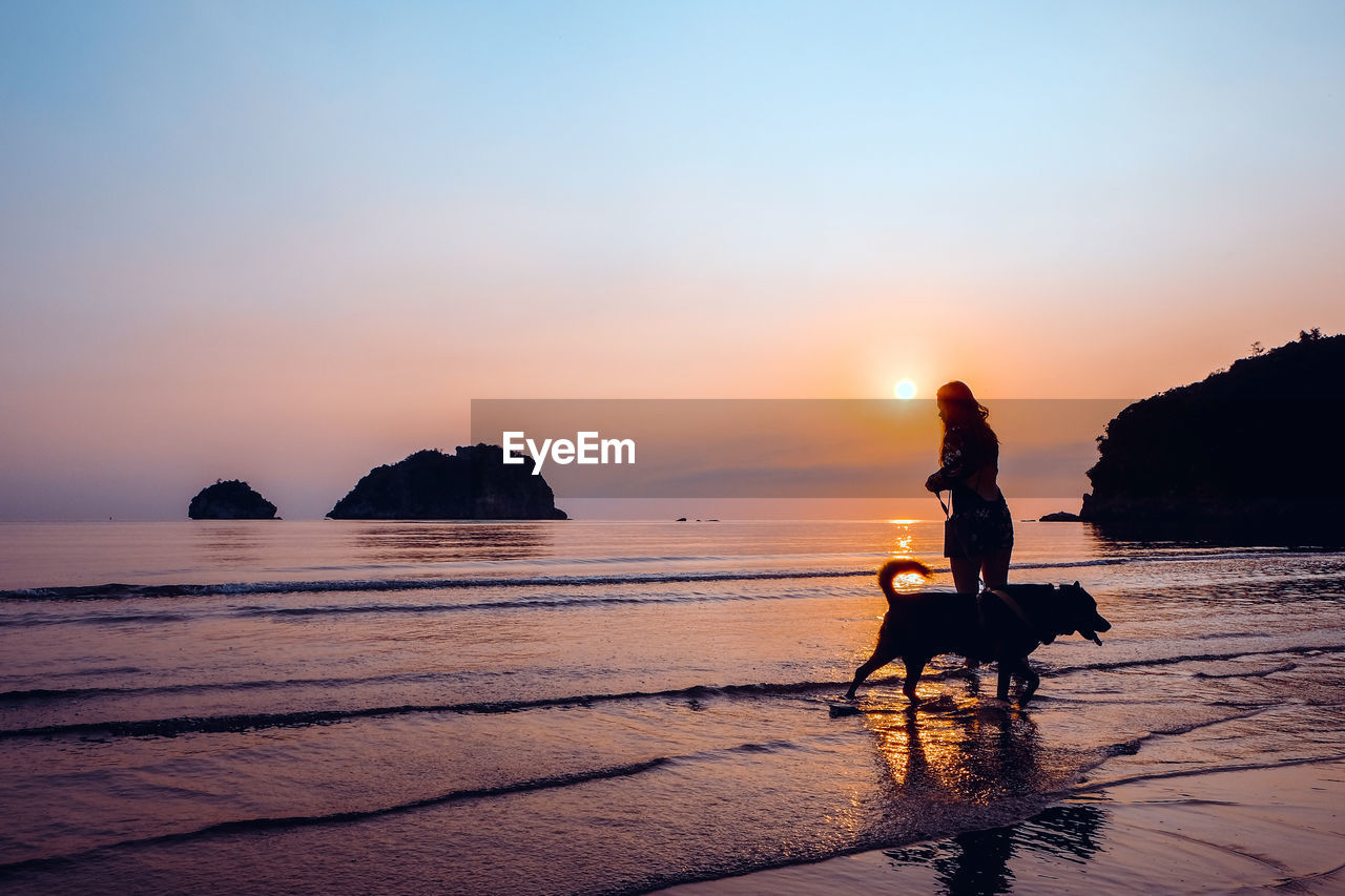 Silhouette mid adult woman with dog standing at beach against sky during sunset