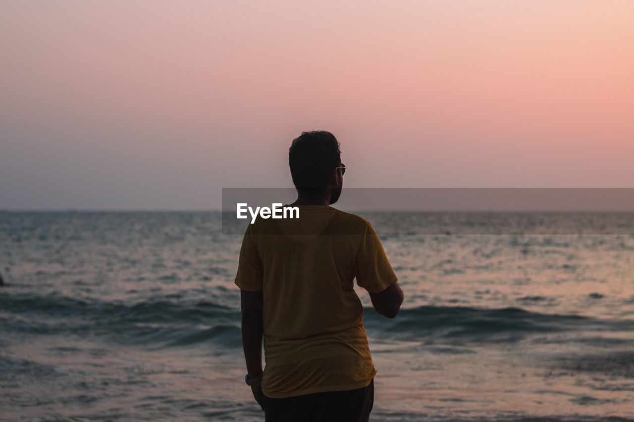 Rear view of man standing at beach against sky during sunset