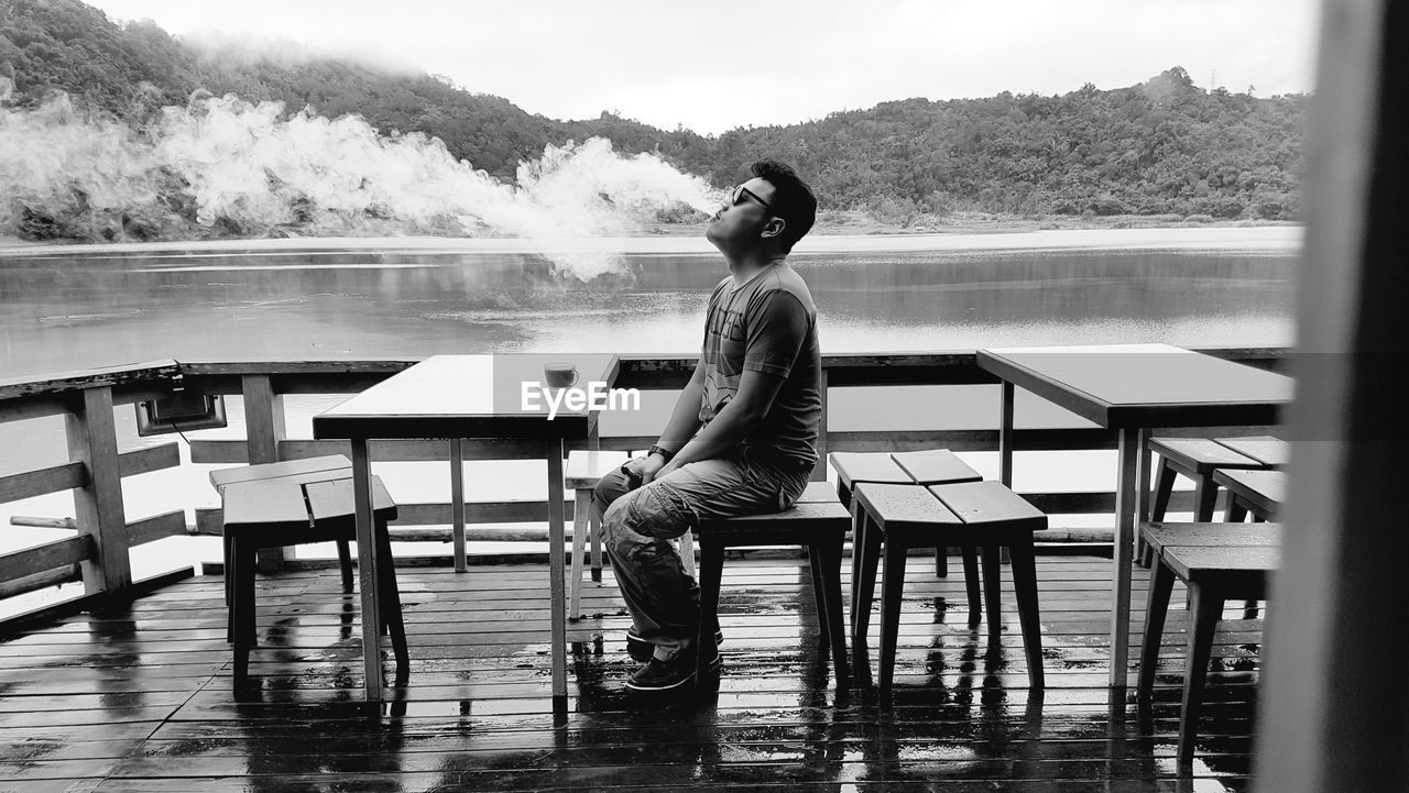 SIDE VIEW OF YOUNG WOMAN SITTING ON TABLE AGAINST CHAIRS AND MOUNTAINS IN WATER