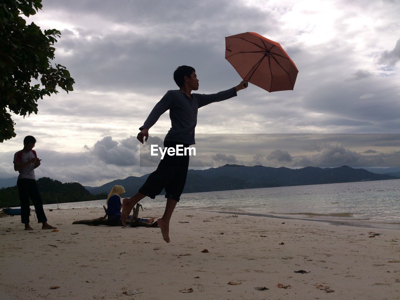Side view of young man with umbrella jumping at beach against cloudy sky
