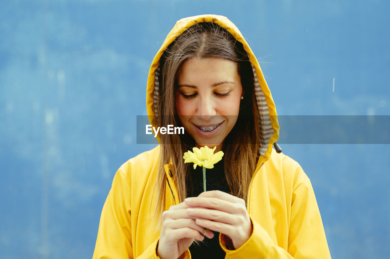 Smiling woman looking at yellow flower standing against blue background