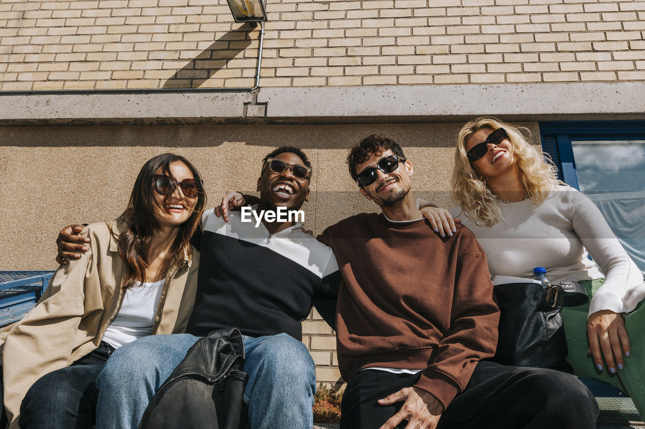 Low angle view of college friends wearing sunglasses and sitting with arms around at college campus