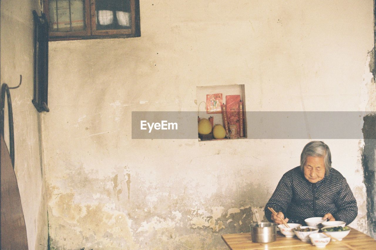 Senior woman eating food while sitting on table against wall