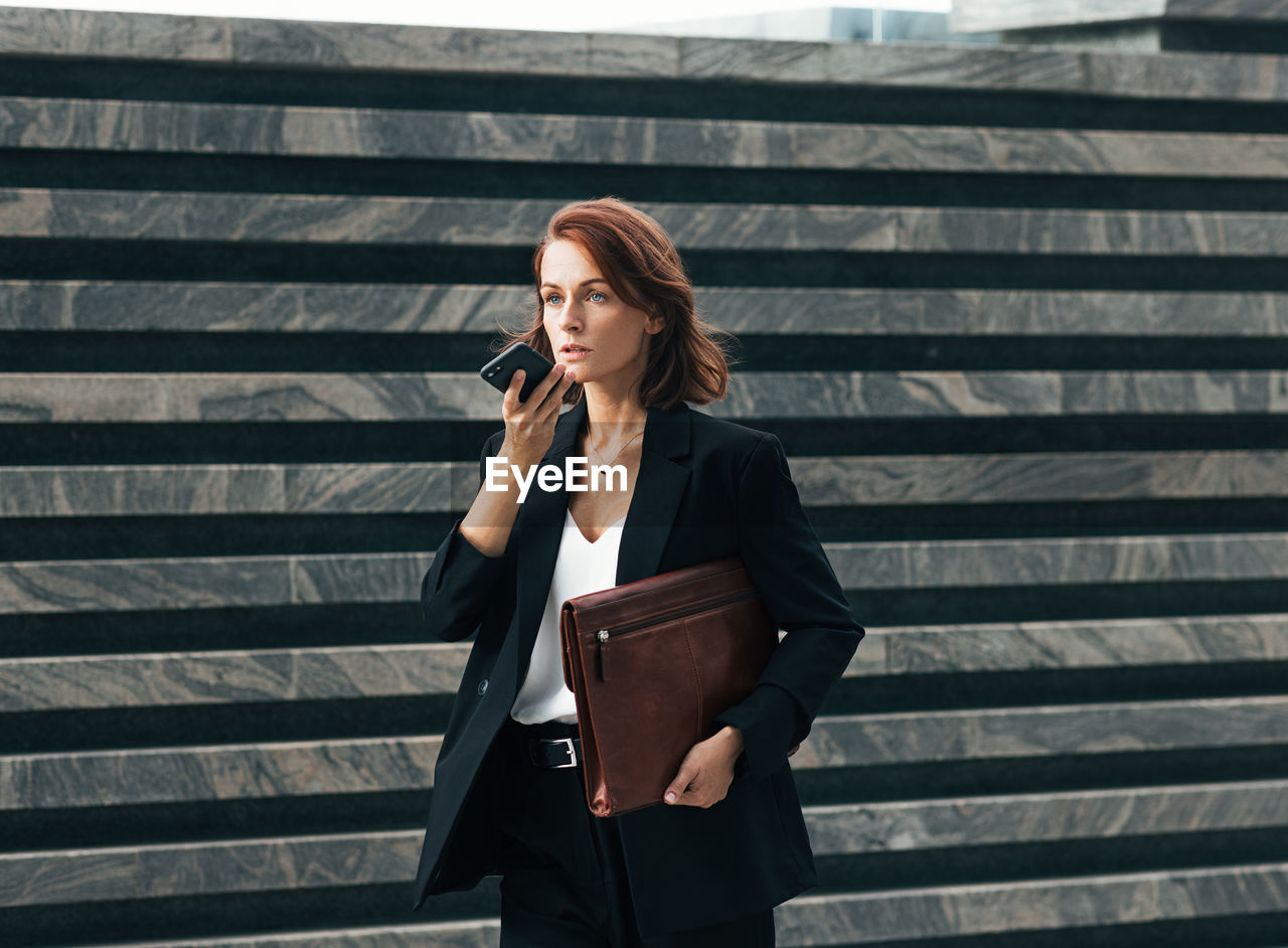 young woman using mobile phone while standing against wall