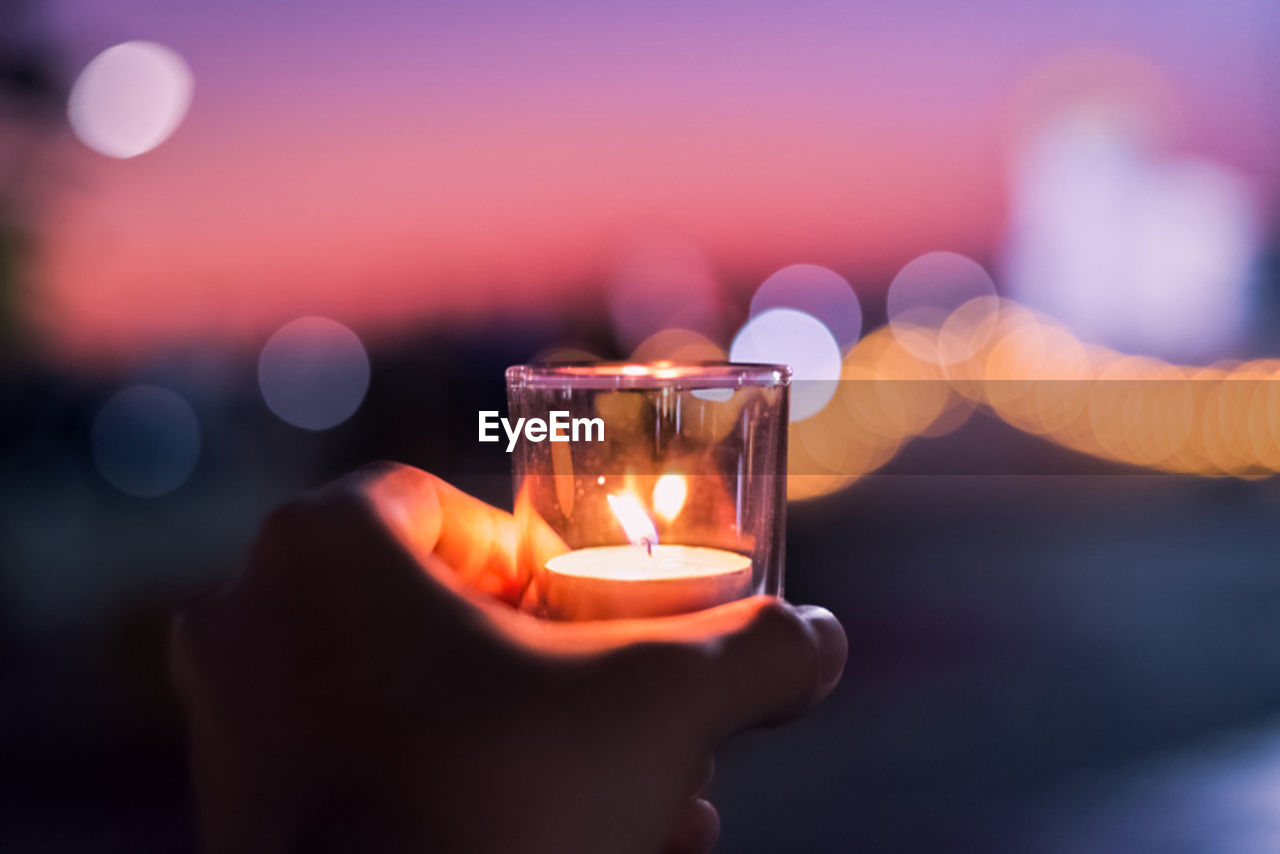 Cropped hand holding lit candle in drinking glass against sky during sunset