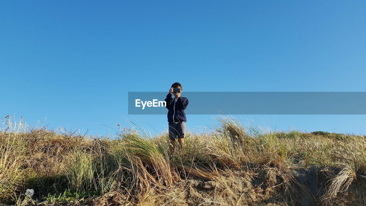 Low angle view of boy photographing with mobile phone against blue sky