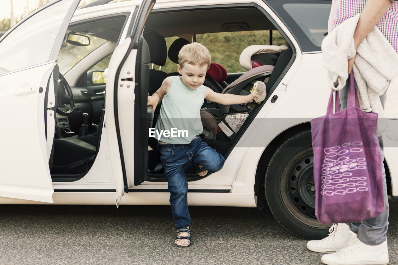 Boy disembarking from car while father standing with bag on street