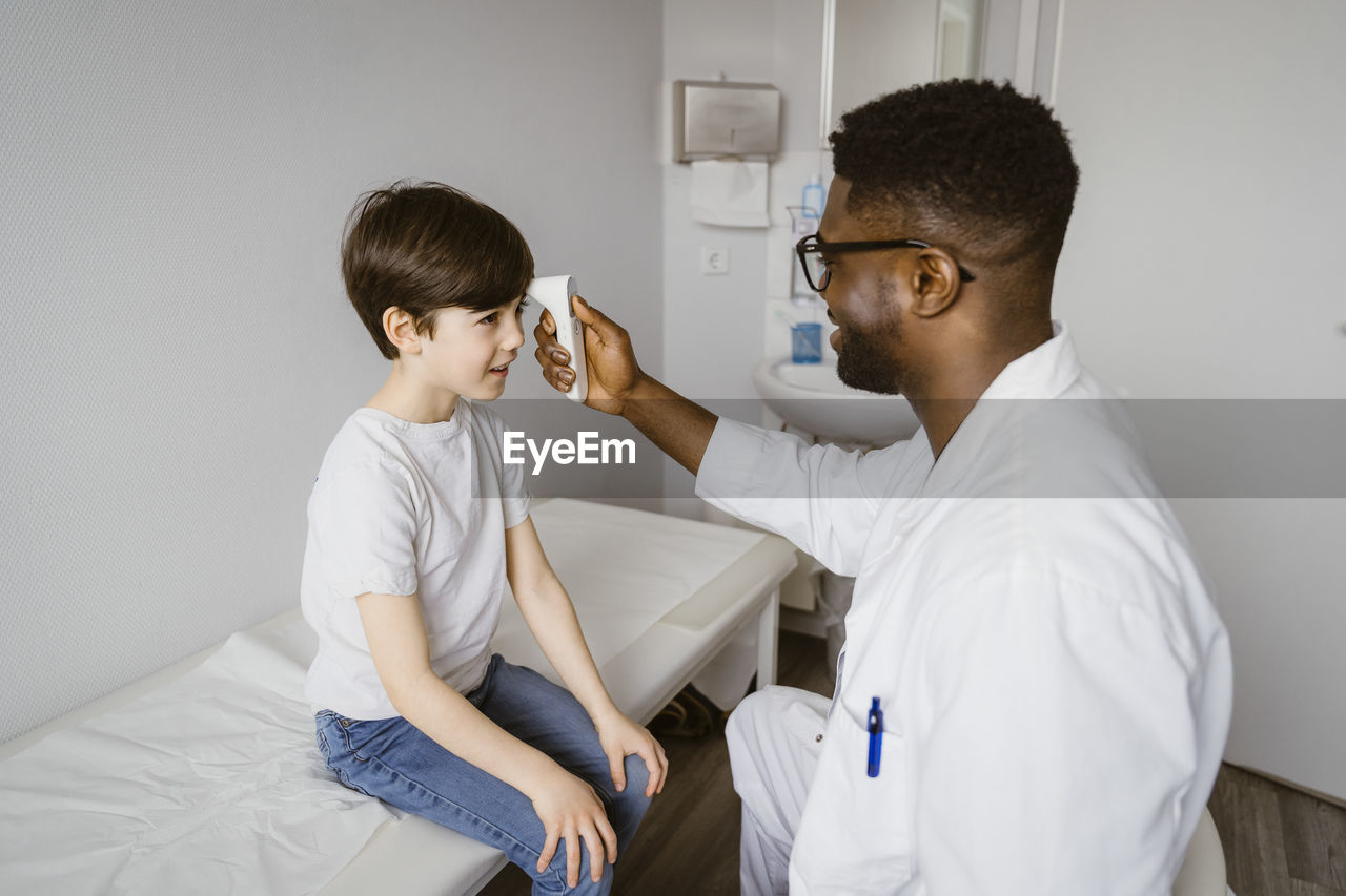 Young male pediatrician measuring temperature of boy using infrared thermometer at clinic