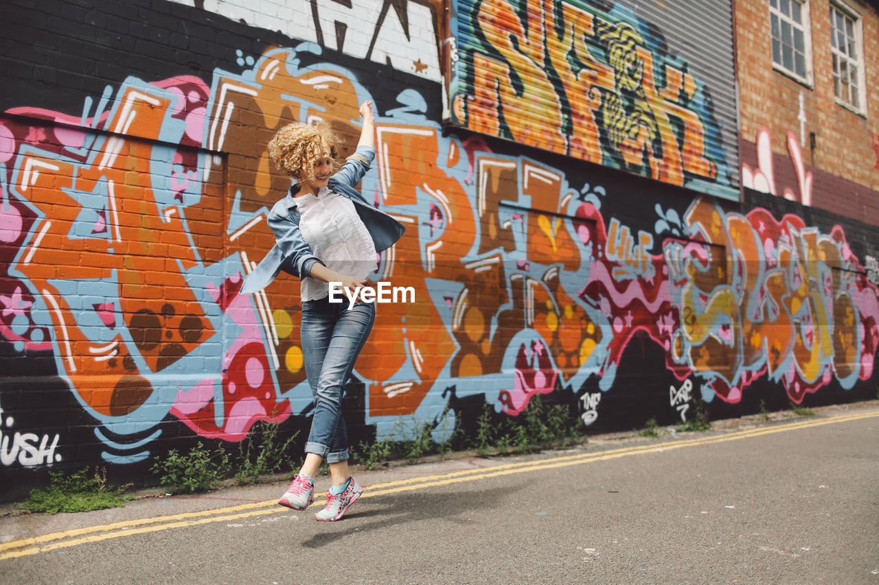 Cheerful woman dancing on road against graffiti wall