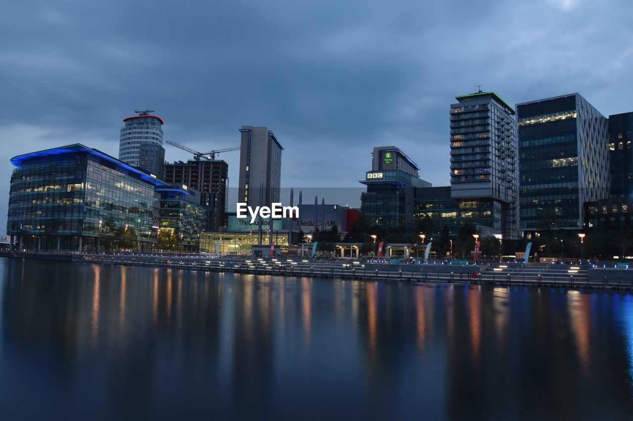 Illuminated buildings by river against sky