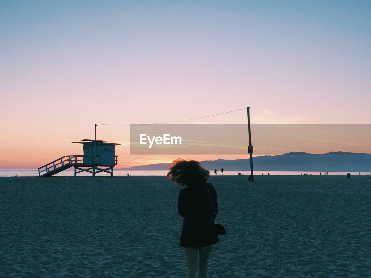 Rear view of woman standing at beach against sky during sunset