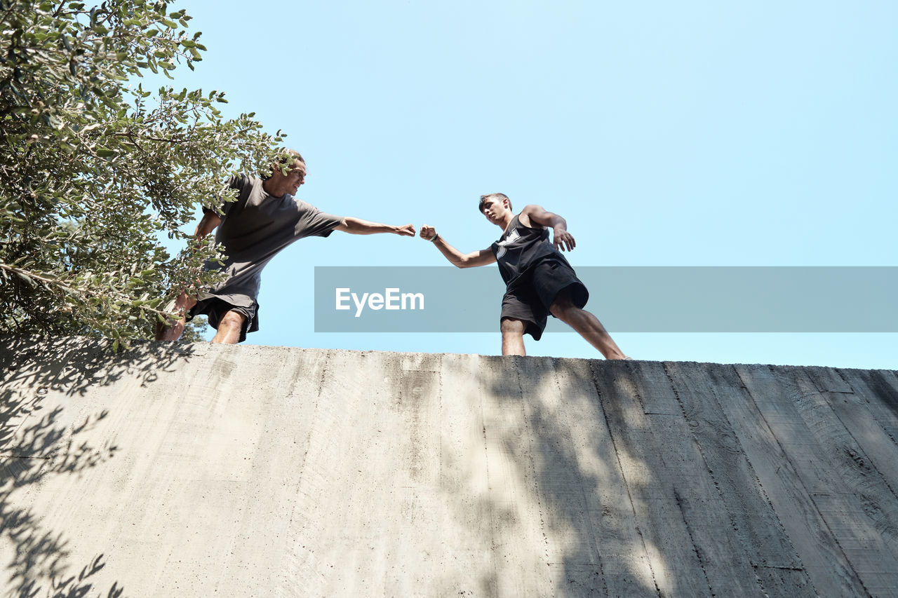 From below of friendly men greeting each other and bumping fists while preparing for parkour training on stone building in city