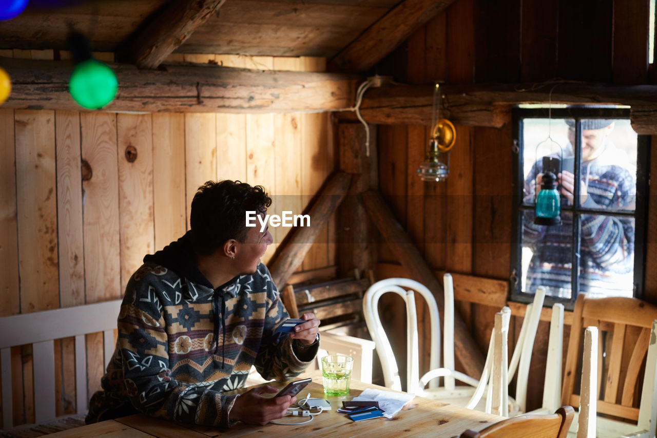 Man photographing friend doing online shopping in cottage from window