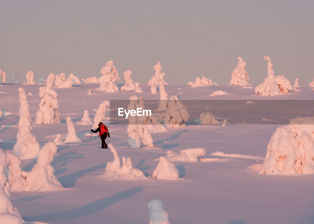 Hiker walking by ice formations on snow covered field against clear sky during winter