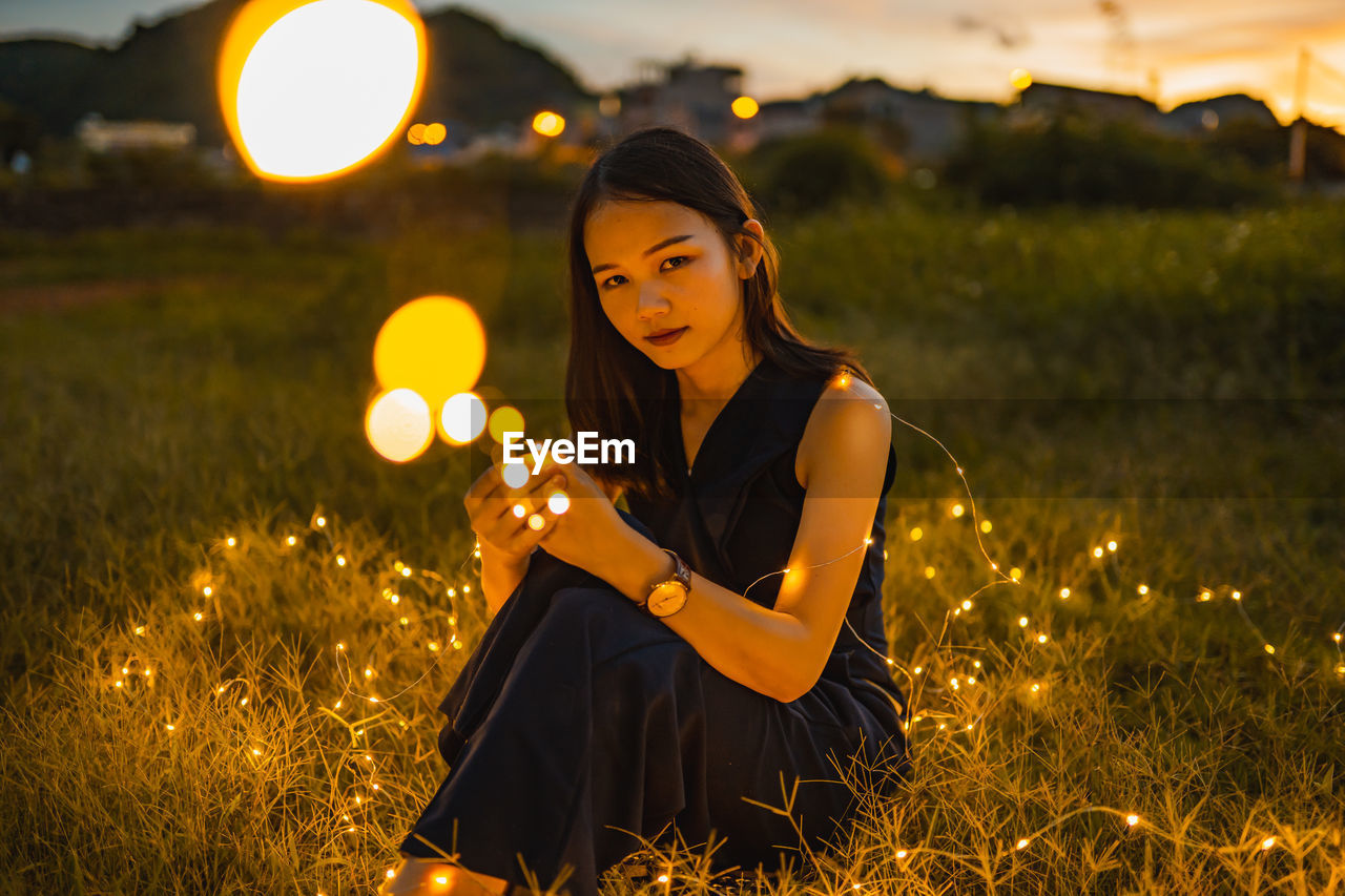 Woman with illuminated string light sitting on grass during sunset