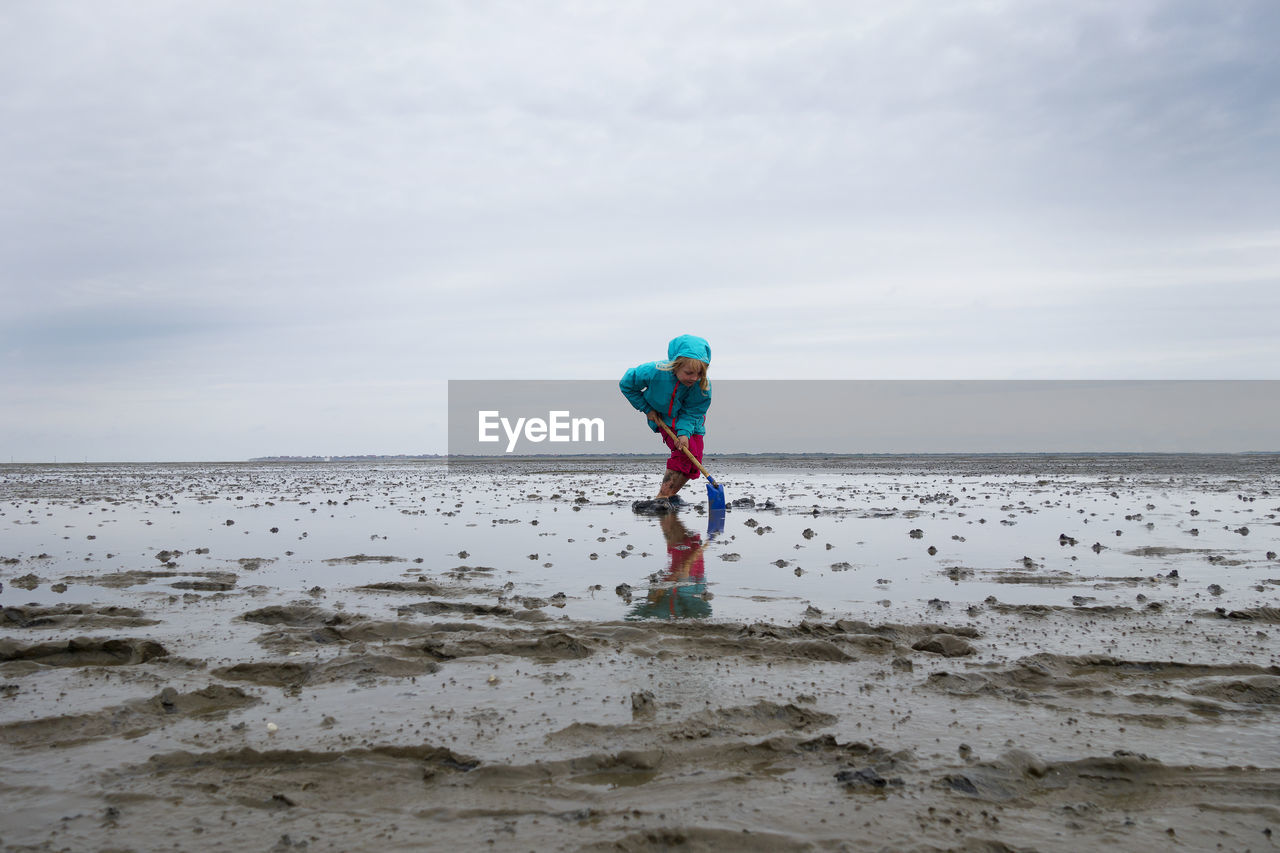 Girl playing in mud flat with shovel against sky at lower saxon wadden sea national park, germany