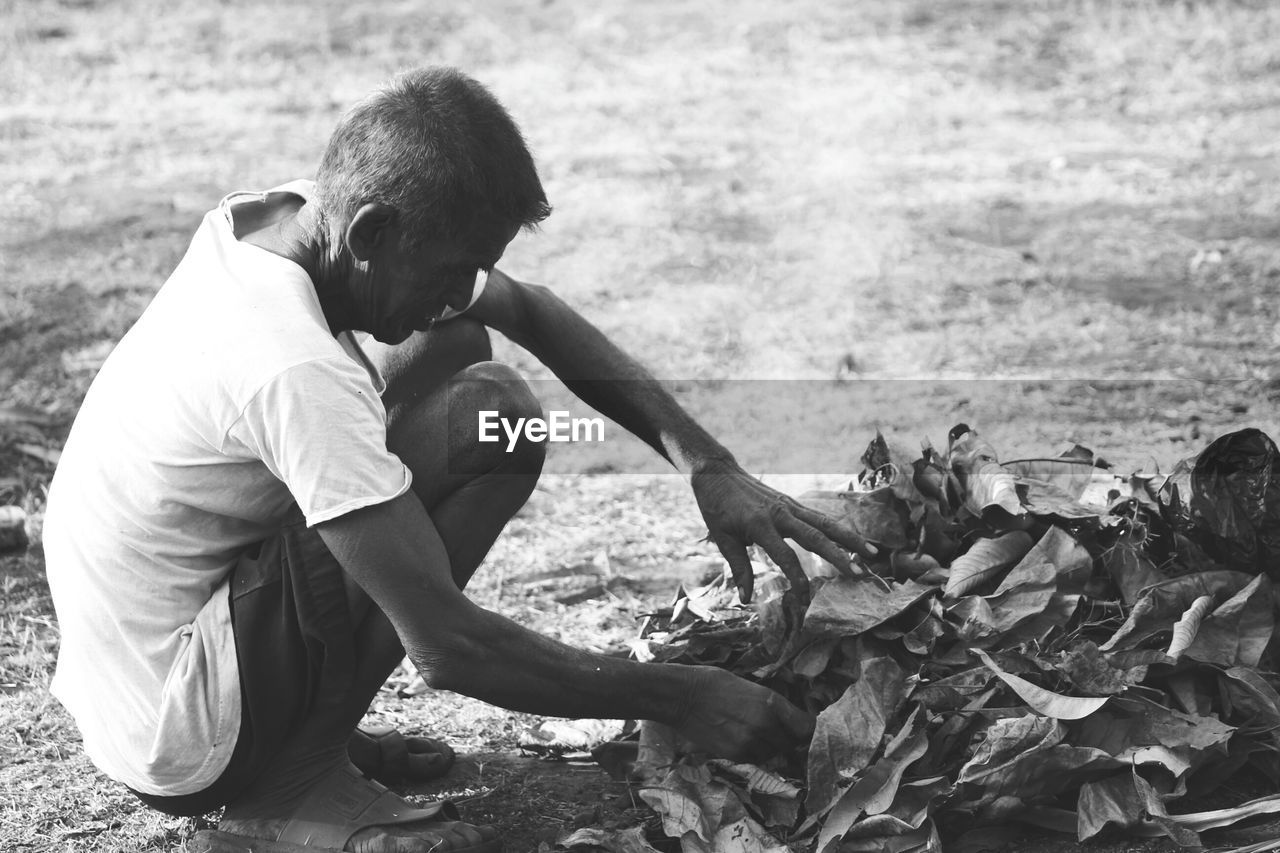 Side view of man crouching on field by heap of leaves