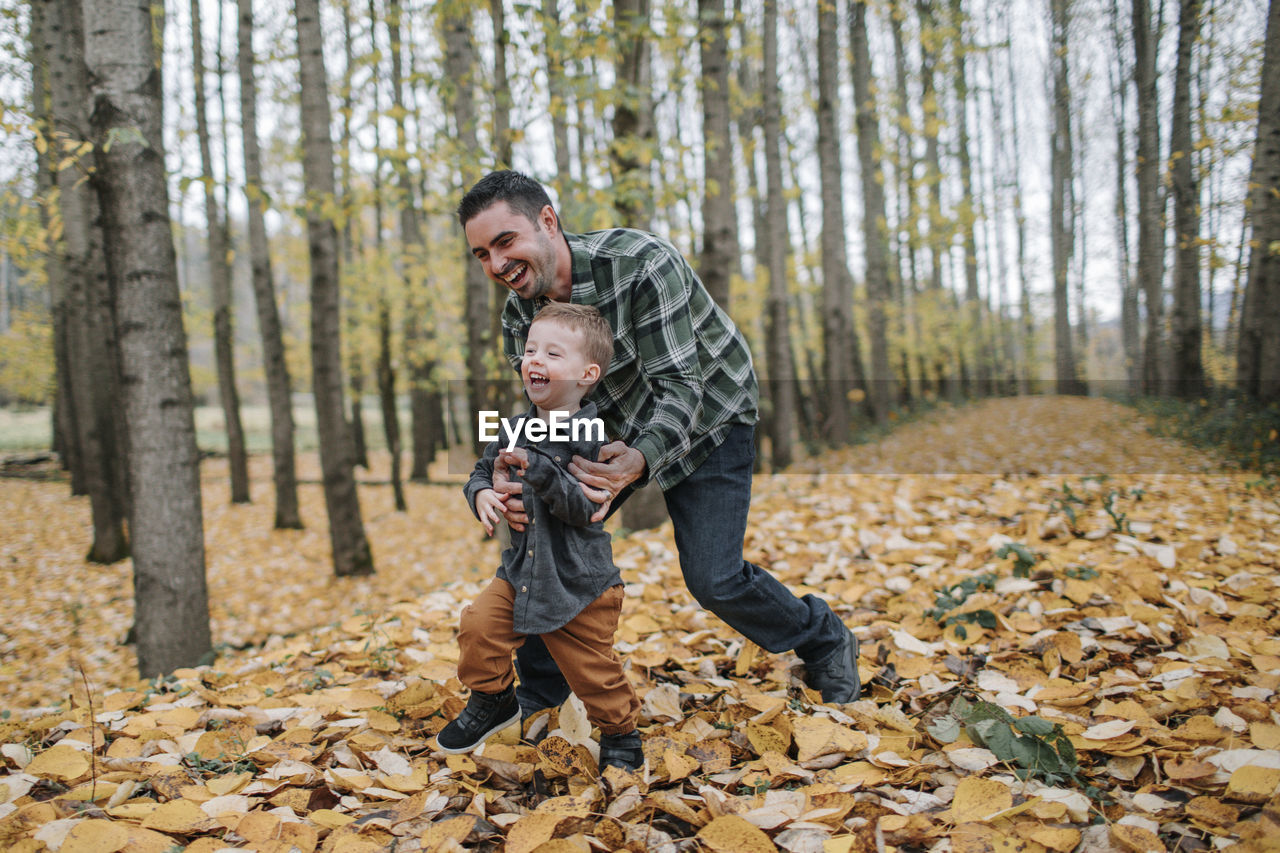 Cheerful father and son playing on dry leaves in forest during autumn