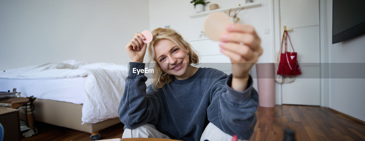portrait of smiling young woman using smart phone while sitting on bed at home