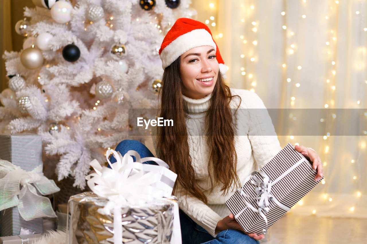 Happy caucasian woman in santa claus hat and light sweater holds a gift box