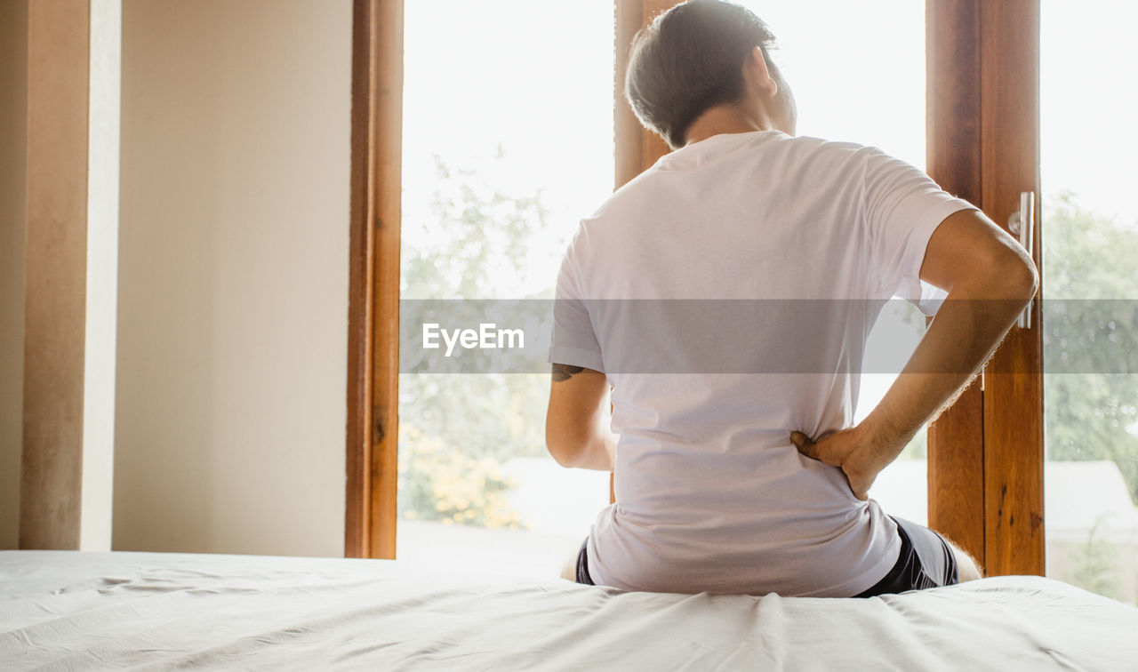 REAR VIEW OF MAN SITTING ON BED